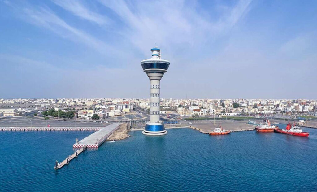 Tall blue-and-white tower rising from a circular base in a harbor, with a city skyline in the distance.
